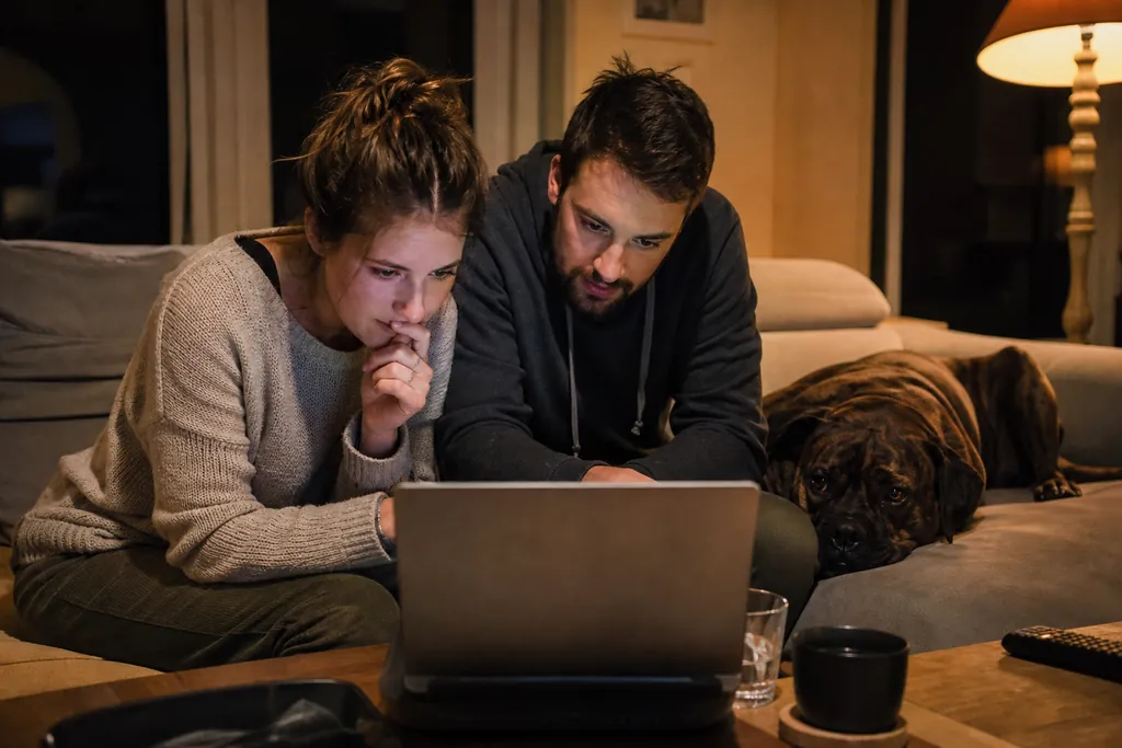 Un couple compare des offres d'assurance sur un ordinateur portable, leur chien la tête posée sur le canapé